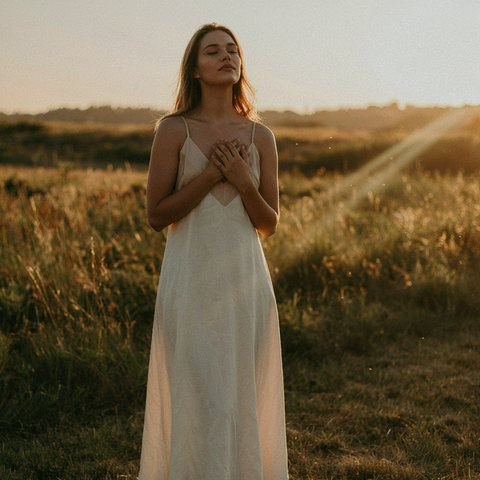 A woman standing in a sunlit field at golden hour, hands resting on her heart
